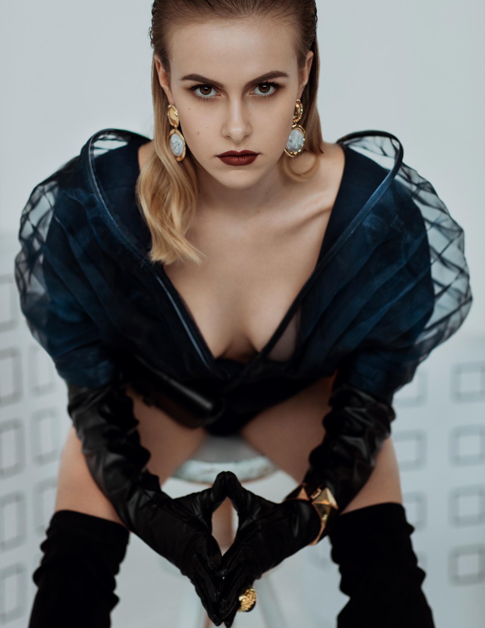 Portret kobiety w czarnych, skórzanych rękawicach i transparentnej bluzce z bufkami, z mocnym makijażem i złotymi kolczykami, siedzącej na białym krześle z wzorzystym tłem.