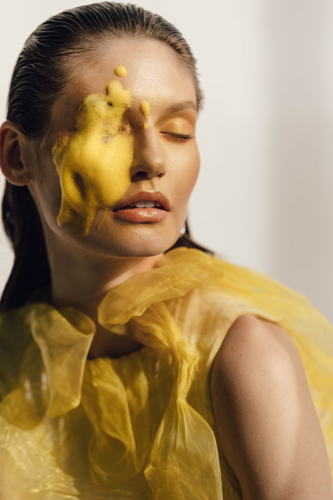 Portret kobiety z zamkniętymi oczami i żółtą substancją na twarzy, ubrana w żółtą, transparentną tkaninę.