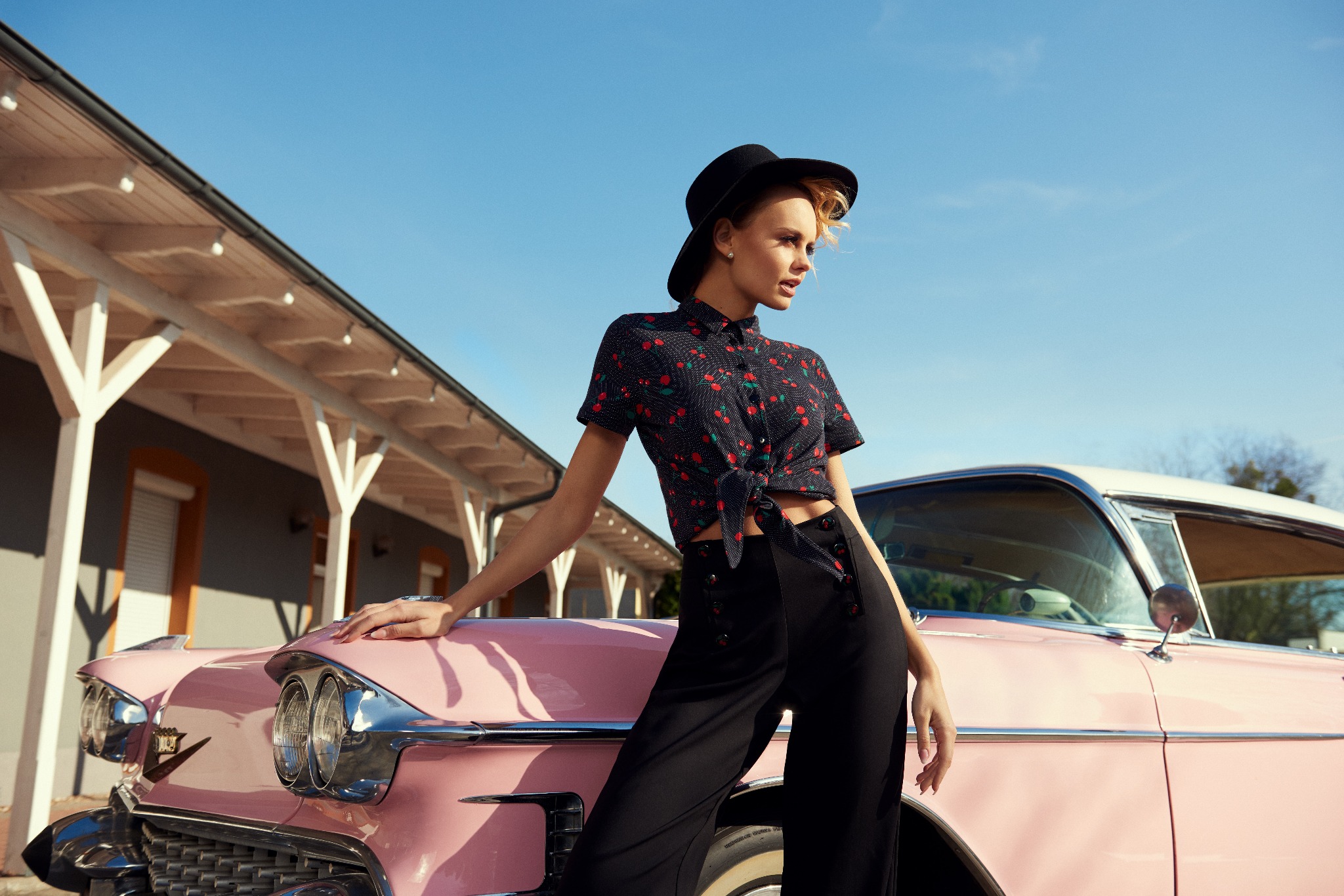 Kobieta w kapeluszu i koszuli w wisienki opiera się o różowy, klasyczny samochód Cadillac na tle budynku z kolumnami.