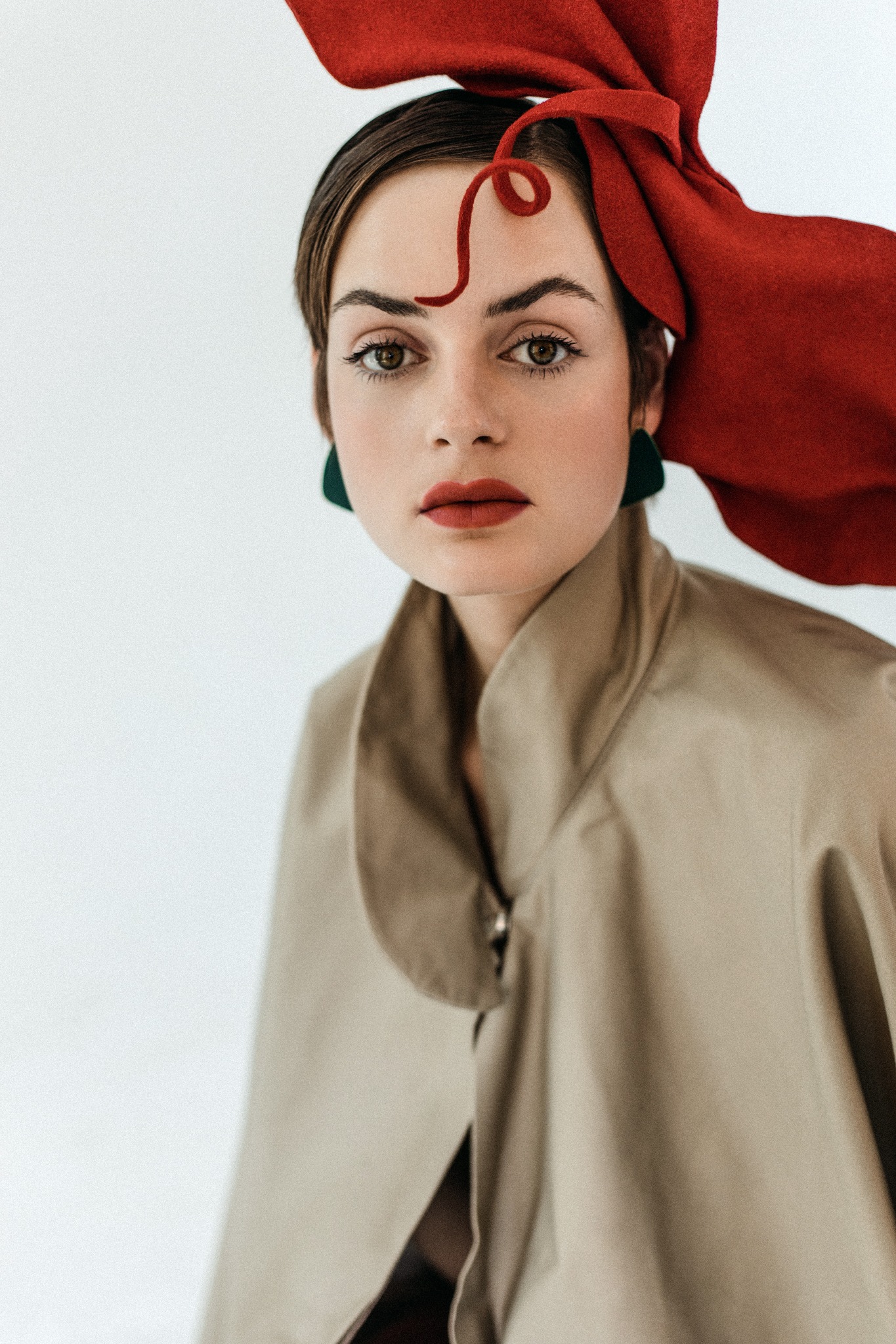 Portret kobiety z czerwonym ozdobnym nakryciem głowy, zielonymi kolczykami i beżowym płaszczem na białym tle.