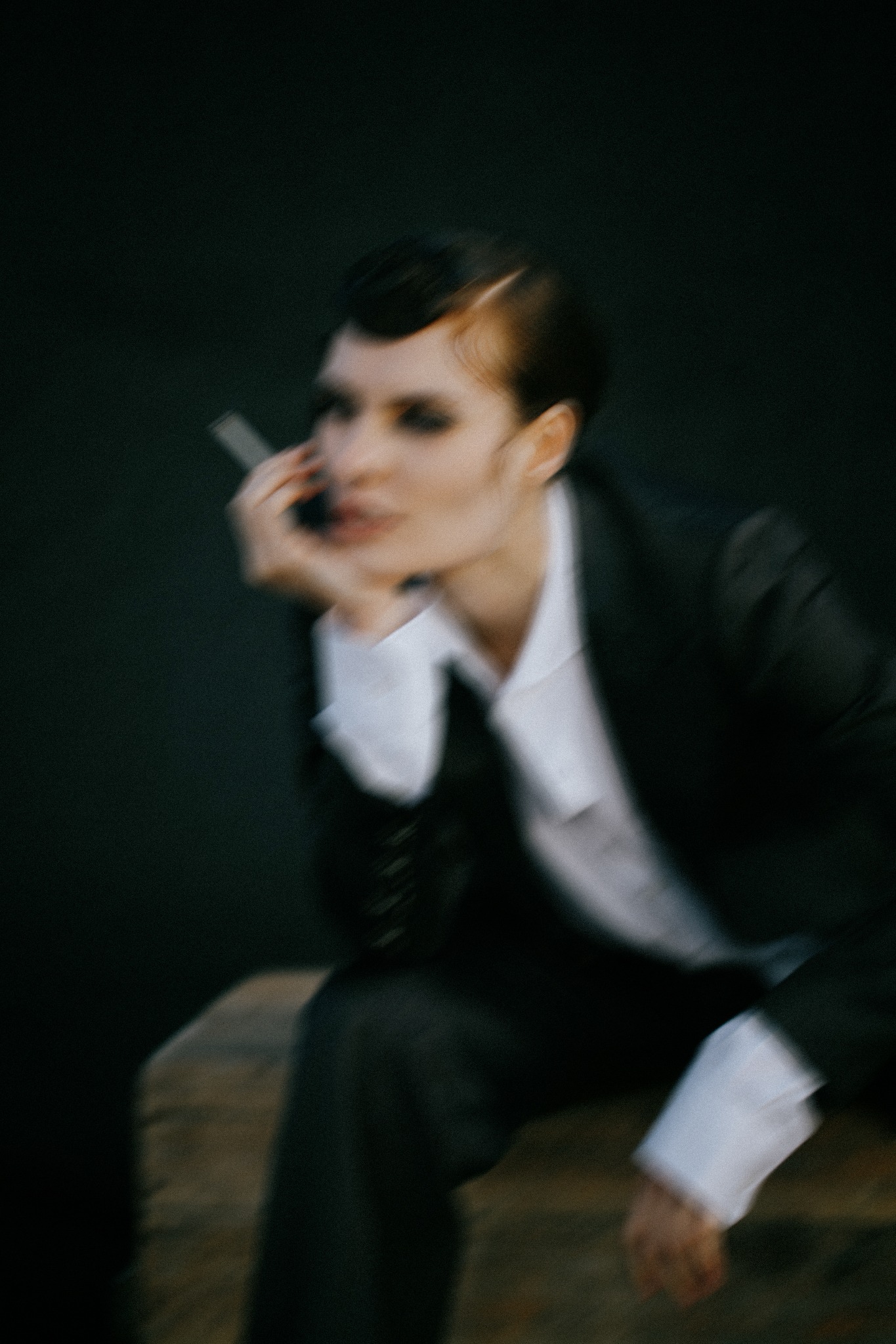 Kobieta w męskim garniturze, z ciemnym makijażem oczu i elegancko zaczesanymi włosami, trzymająca papierosa przy twarzy, w rozmyciu.