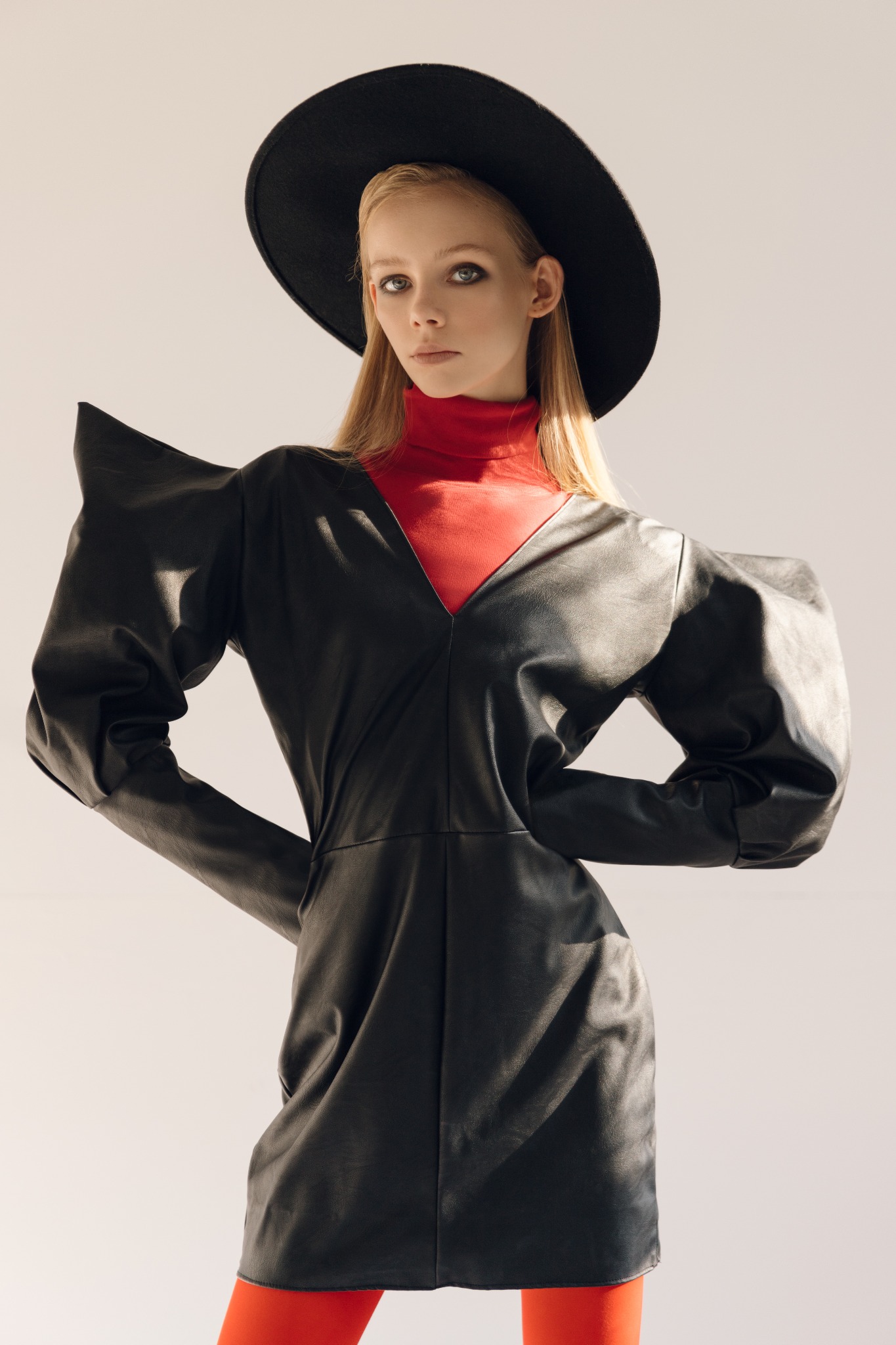Modelka w czarnej, skórzanej sukience z bufkami, czerwonym golfie i czarnym kapeluszu na tle jasnego studia.