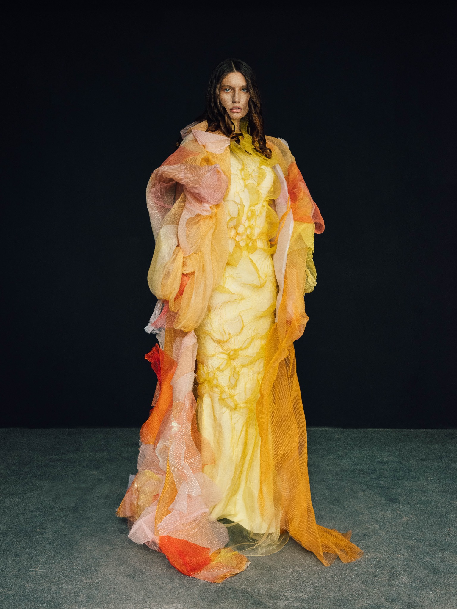 Modelka w żółtej sukni z warstwami pomarańczowego, różowego i białego tiulu, na ciemnym tle.