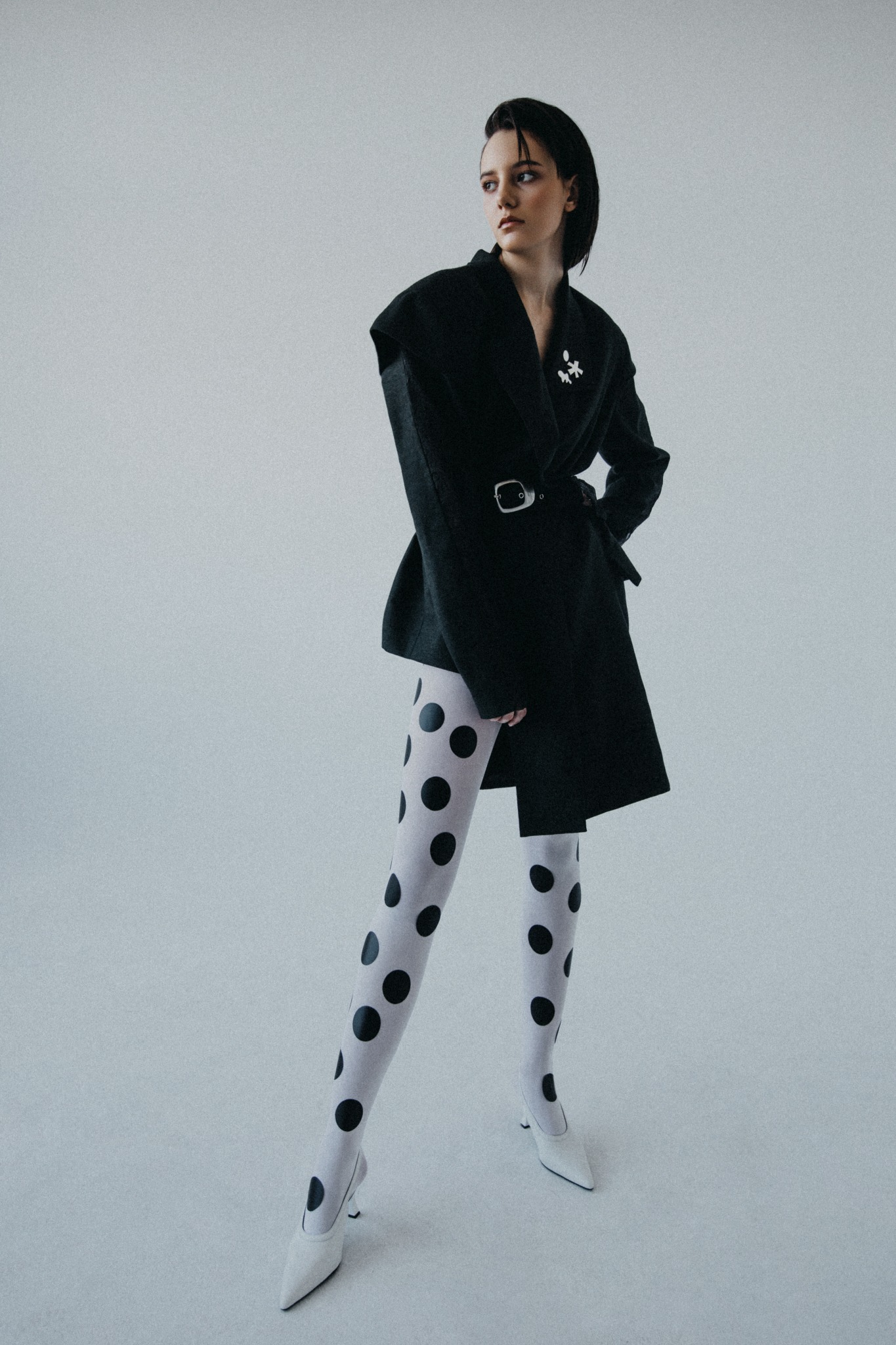 Kobieta w czarnej oversize marynarce z paskiem, białych rajstopach w czarne kropki i białych szpilkach na jednolitym, jasnym tle. Marynarka ozdobiona broszką.