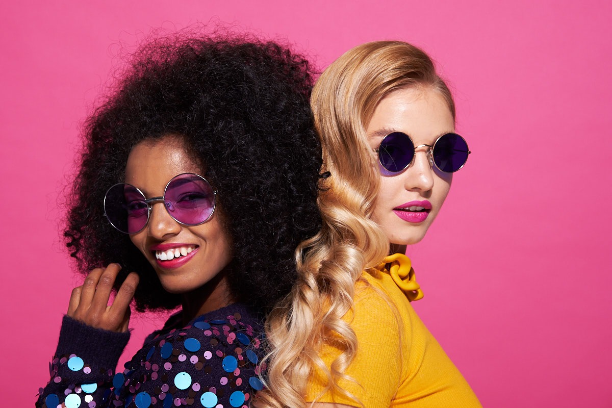 Dwie uśmiechnięte kobiety o różnej karnacji i fryzurach, w okrągłych okularach przeciwsłonecznych, pozują na tle różowego studia.