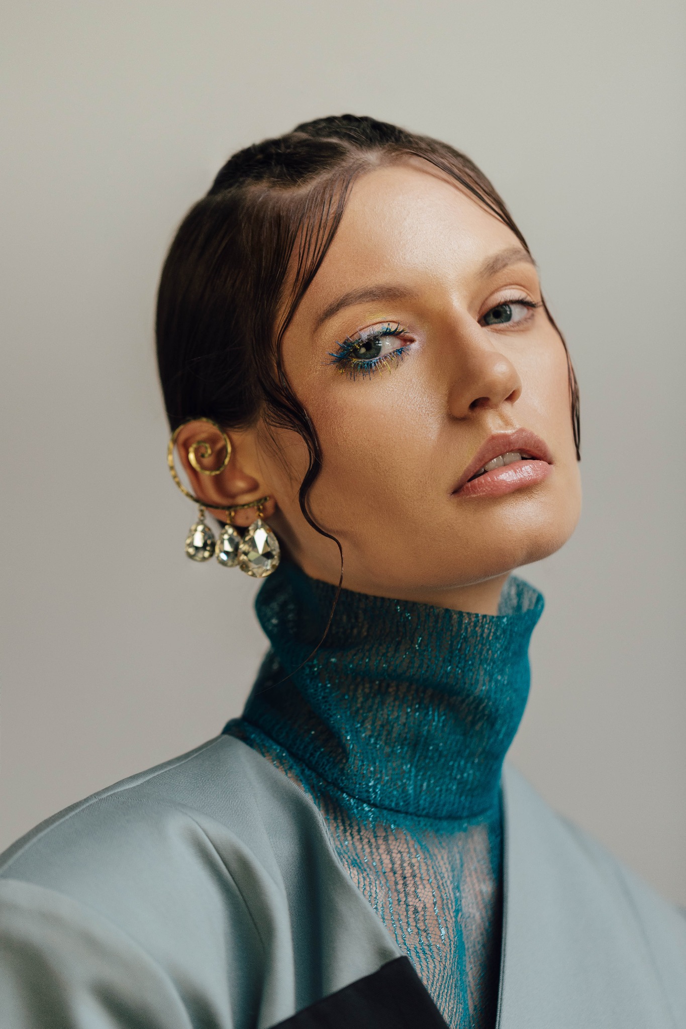 Portret kobiety z awangardowym makijażem oczu w odcieniach żółci i błękitu, błyszczącymi ustami oraz ozdobnym kolczykiem oplatającym ucho, ubrana w turkusowy golf i jasnoszarą marynarkę.