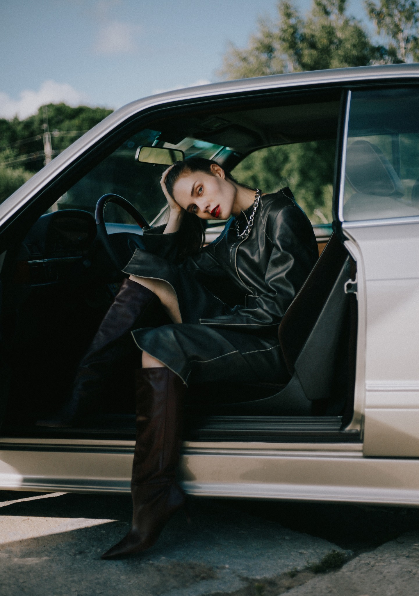 Kobieta z intensywnym makijażem w czarnej, skórzanej kurtce i wysokich, brązowych kozakach pozuje w srebrnym samochodzie z otwartymi drzwiami.