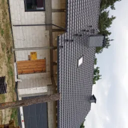 Dom w trakcie budowy z częściowo ukończonym, czarnym dachem z oknem dachowym i kominem, widoczne elementy konstrukcyjne i materiały budowlane.