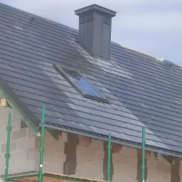 Nowy dach z szarej dachówki z oknem dachowym i kominem wentylacyjnym, widok z rusztowania na budowie.