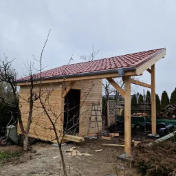 Drewniana altana w trakcie budowy z czerwonym dachem, widoczna drabina, przyczepa z oponami i sterta drewna na tle pochmurnego nieba.