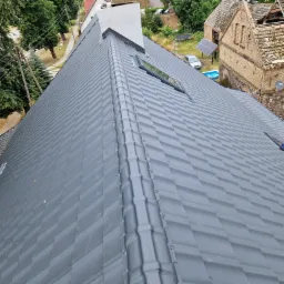 Nowy, grafitowy dach z blachy trapezowej z oknem dachowym i kominem, widok z góry na tle zieleni i zabudowań.