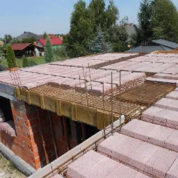 Wznoszenie ścian domu z czerwonej cegły, widoczne zbrojenie i szalunek stropu w trakcie budowy, w tle zabudowa jednorodzinna.