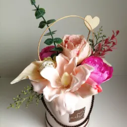 Kompozycja kwiatowa w okrągłym, jasnoróżowym pudełku z napisem 'Flower' i sercem, z drewnianym serduszkiem na patyku, różowe i kremowe kwiaty, gałązki eukaliptusa i dekoracyjna kula z wikliny.