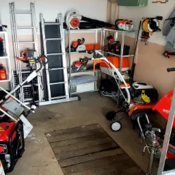 Magazyn z narzędziami ogrodniczymi: kosiarki, pilarki, dmuchawy, opryskiwacz, glebogryzarka, drabina i inne akcesoria na metalowych regałach w garażu.