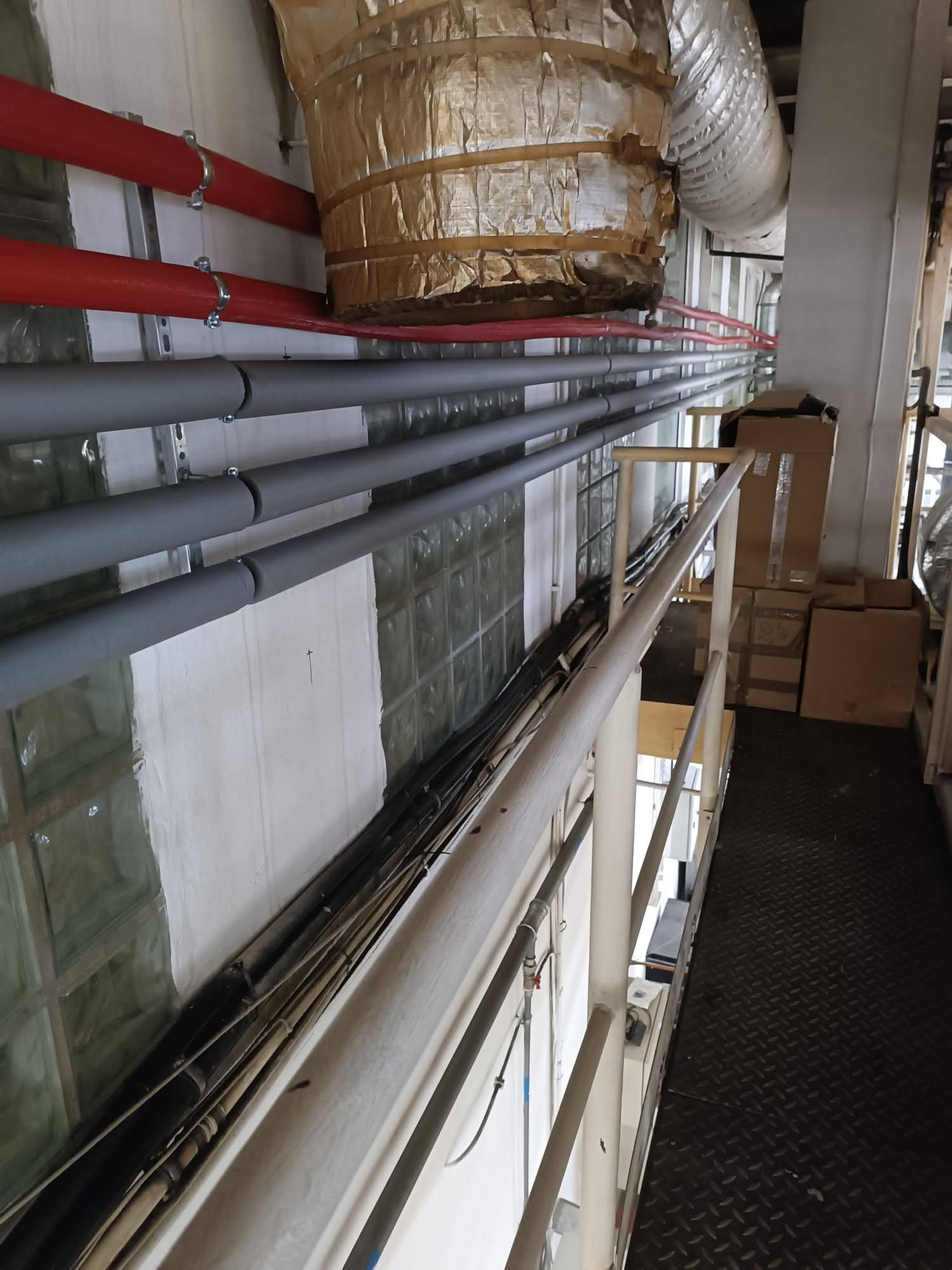 Różne rury instalacyjne (czerwone, szare, czarne) prowadzone wzdłuż ściany z luksferami, zabezpieczone uchwytami, z widocznym fragmentem metalowej konstrukcji i izolacją termiczną rury wentylacyjnej.