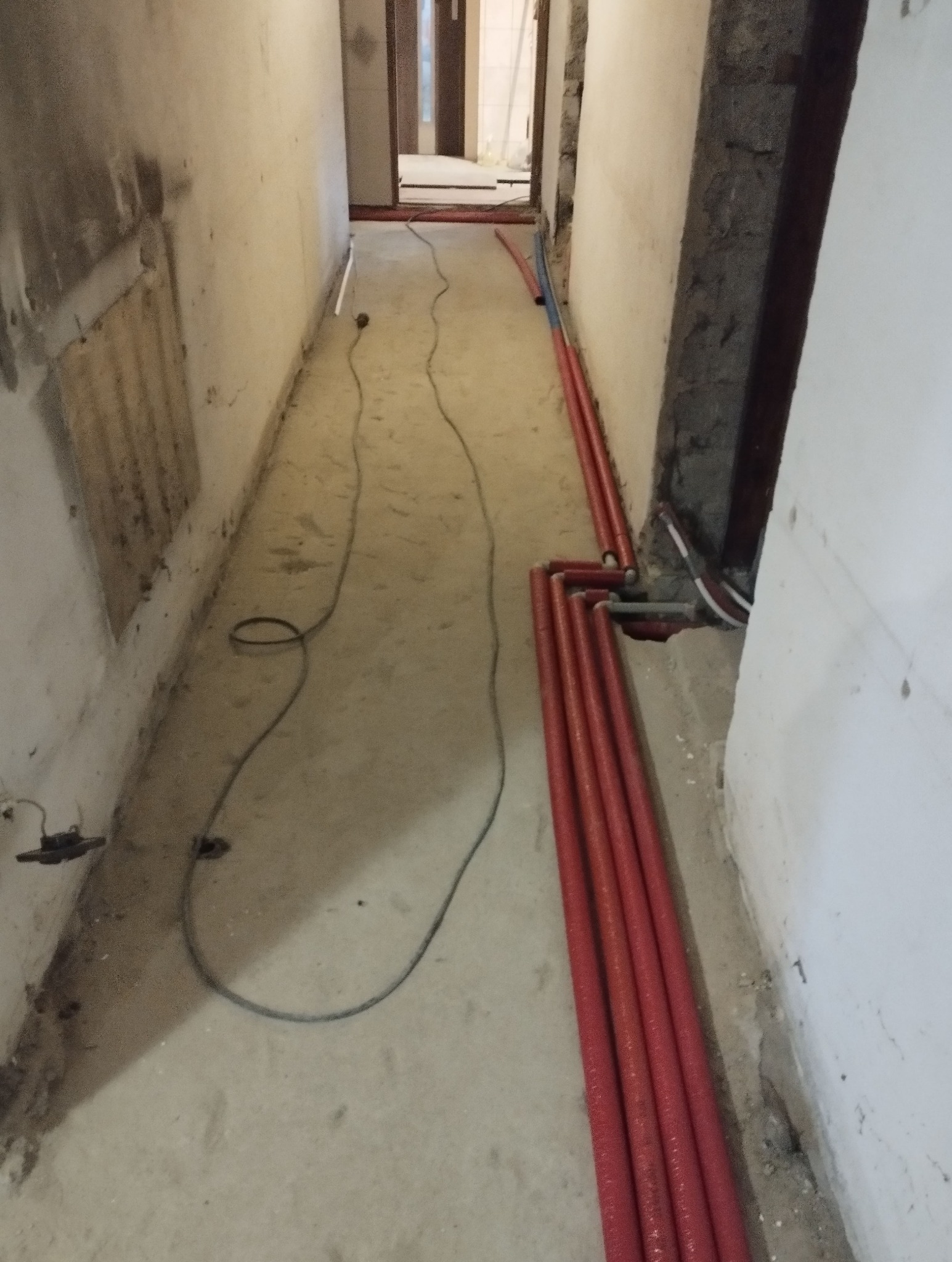Korytarz w trakcie remontu z ułożonymi czerwonymi rurami grzewczymi przy ścianie i wystającym kablem elektrycznym na podłodze.