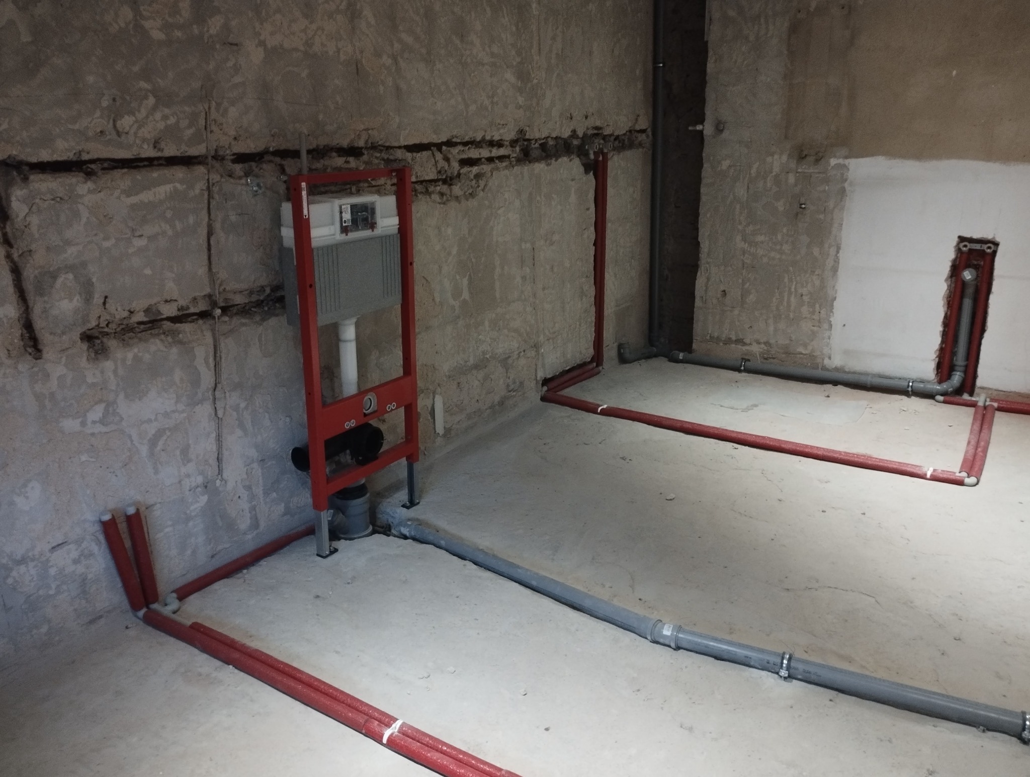 Instalacja wodno-kanalizacyjna w trakcie montażu, widoczne rury czerwone i szare, stelaż podtynkowy WC na tle surowej ściany.