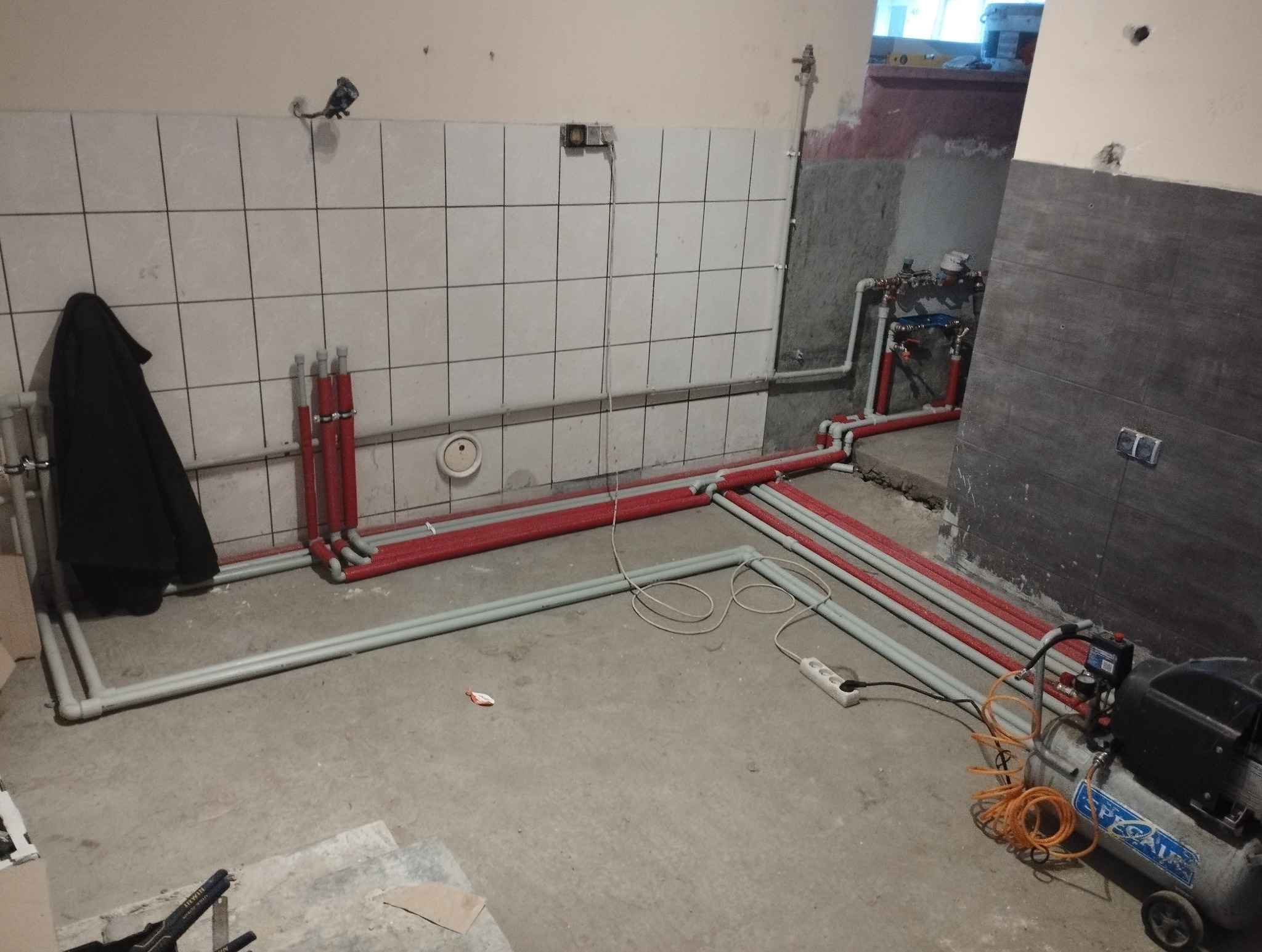 Wnętrze w trakcie prac hydraulicznych z widocznymi rurami instalacji wodnej i grzewczej, kompresorem oraz fragmentem czarnego płaszcza wiszącego na ścianie z białymi płytkami.