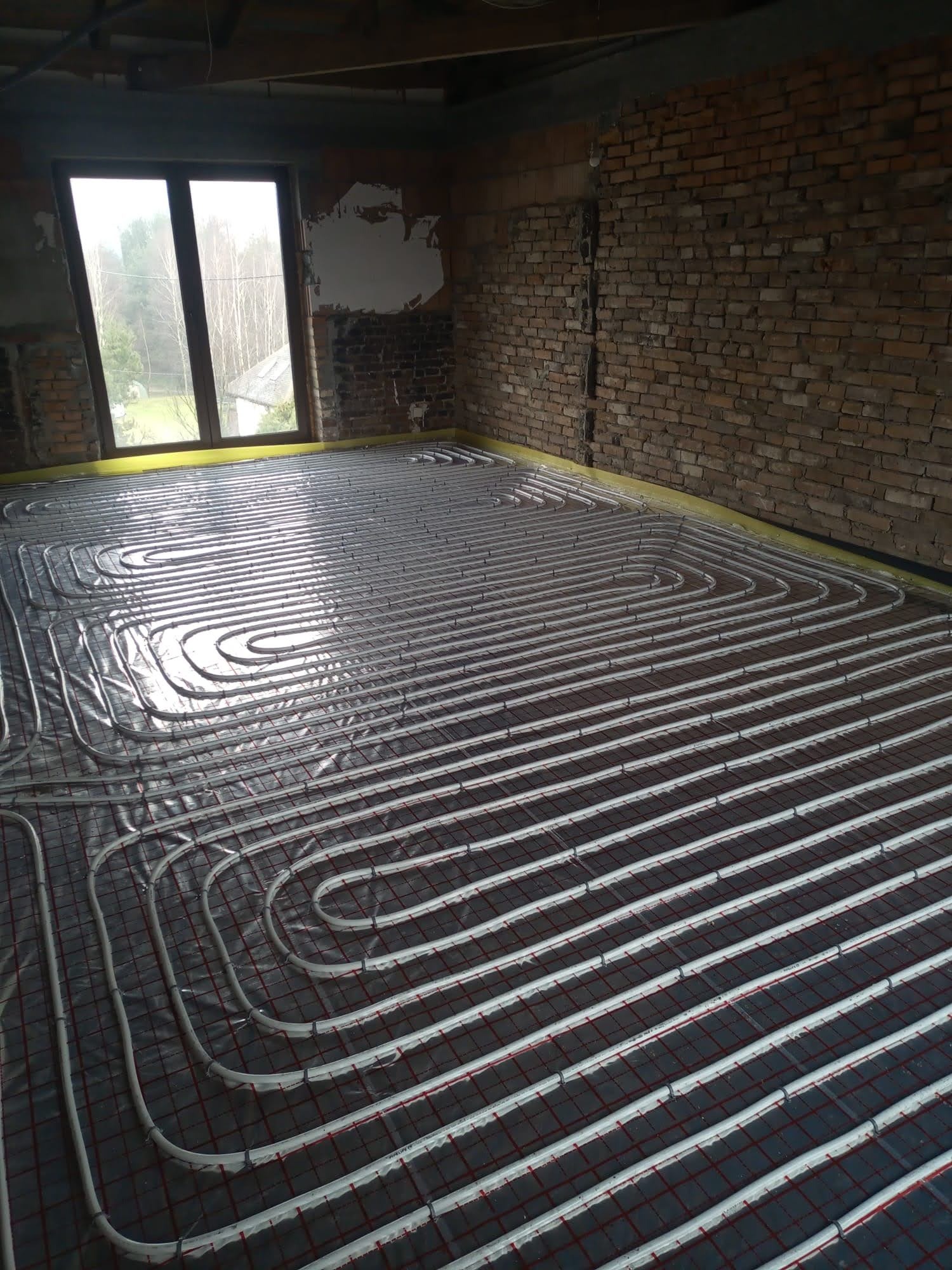 Montaż rur ogrzewania podłogowego na siatce w surowym wnętrzu z ceglaną ścianą w Świerklanach, widoczne okno z widokiem na drzewa.