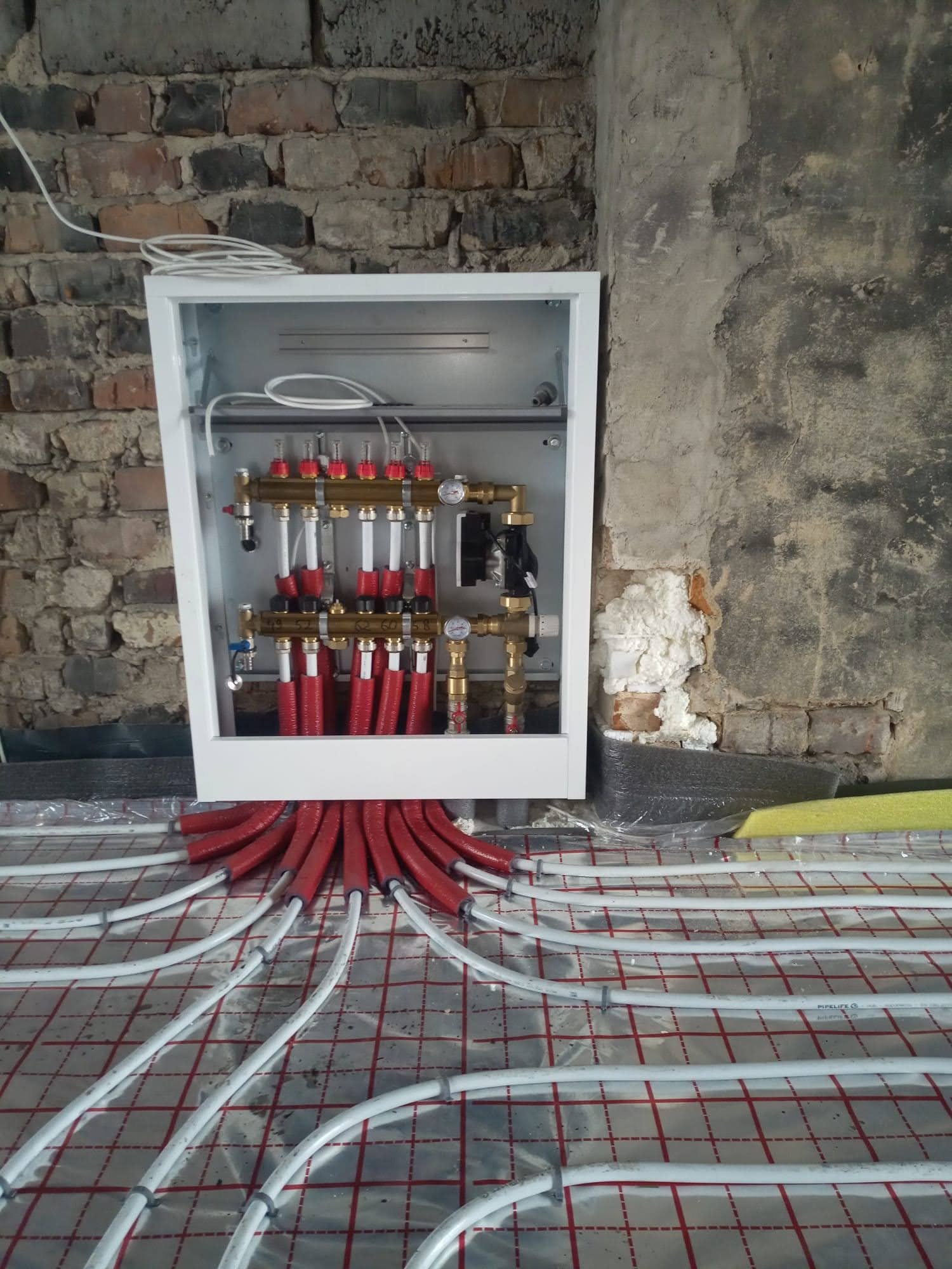 Rozdzielacz ogrzewania podłogowego w białej obudowie, z czerwonymi rurami wychodzącymi do białych przewodów na folii z czerwoną siatką, na tle surowej ściany z cegły i betonu.