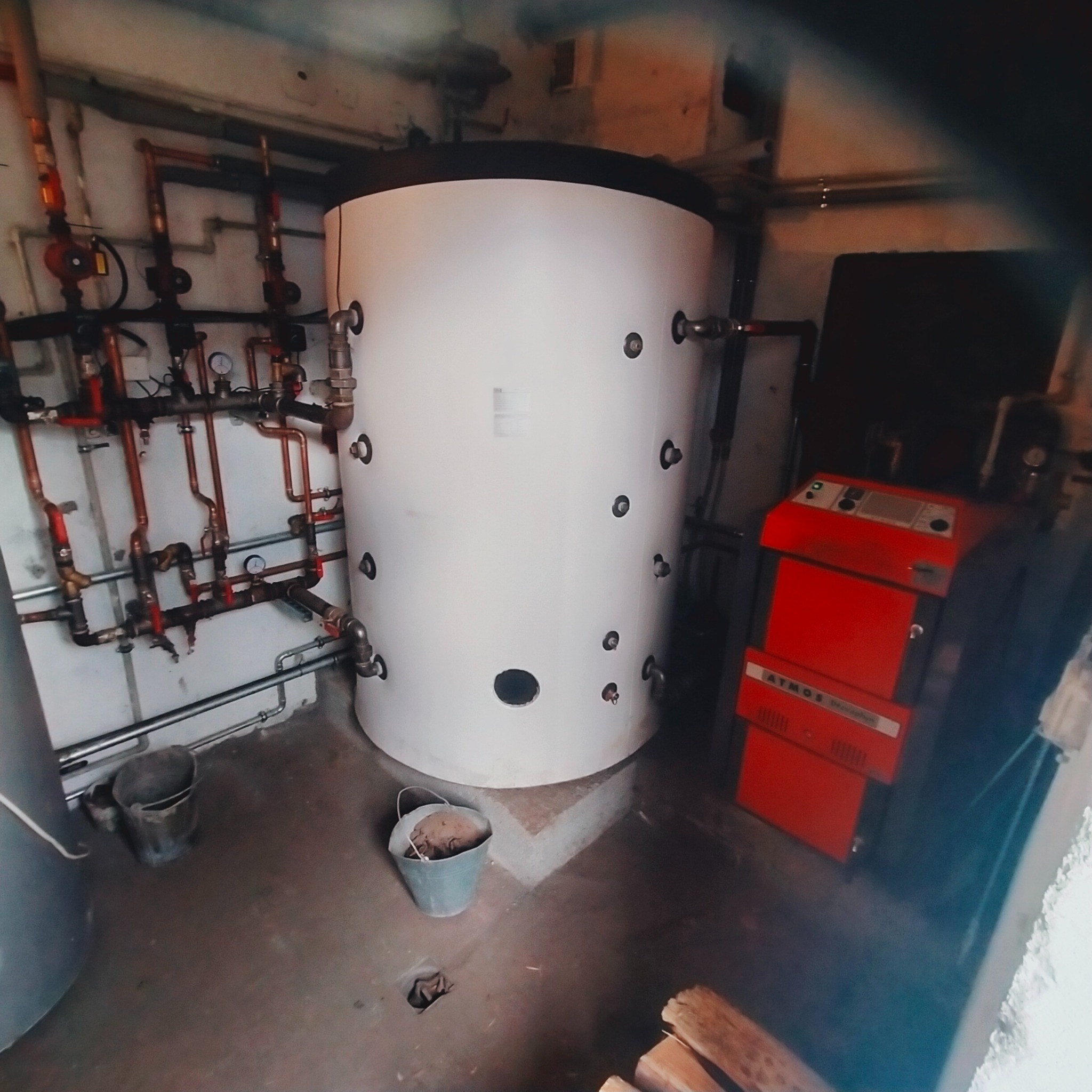 Biały akumulacyjny zbiornik ciepłej wody z licznymi przyłączami, obok czerwony piec Atmos, widoczne rury instalacji grzewczej i dwa wiadra na podłodze w piwnicy.