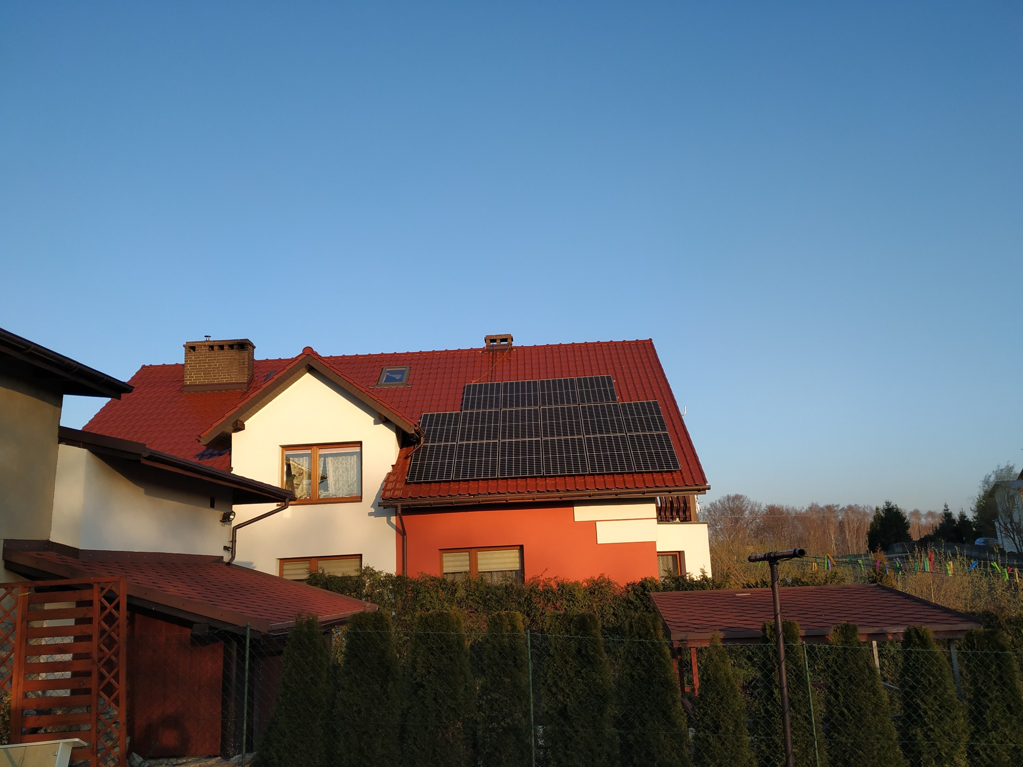 Dom jednorodzinny z czerwoną dachówką i panelami fotowoltaicznymi na dachu, widoczne zza żywopłotu w słoneczny dzień.