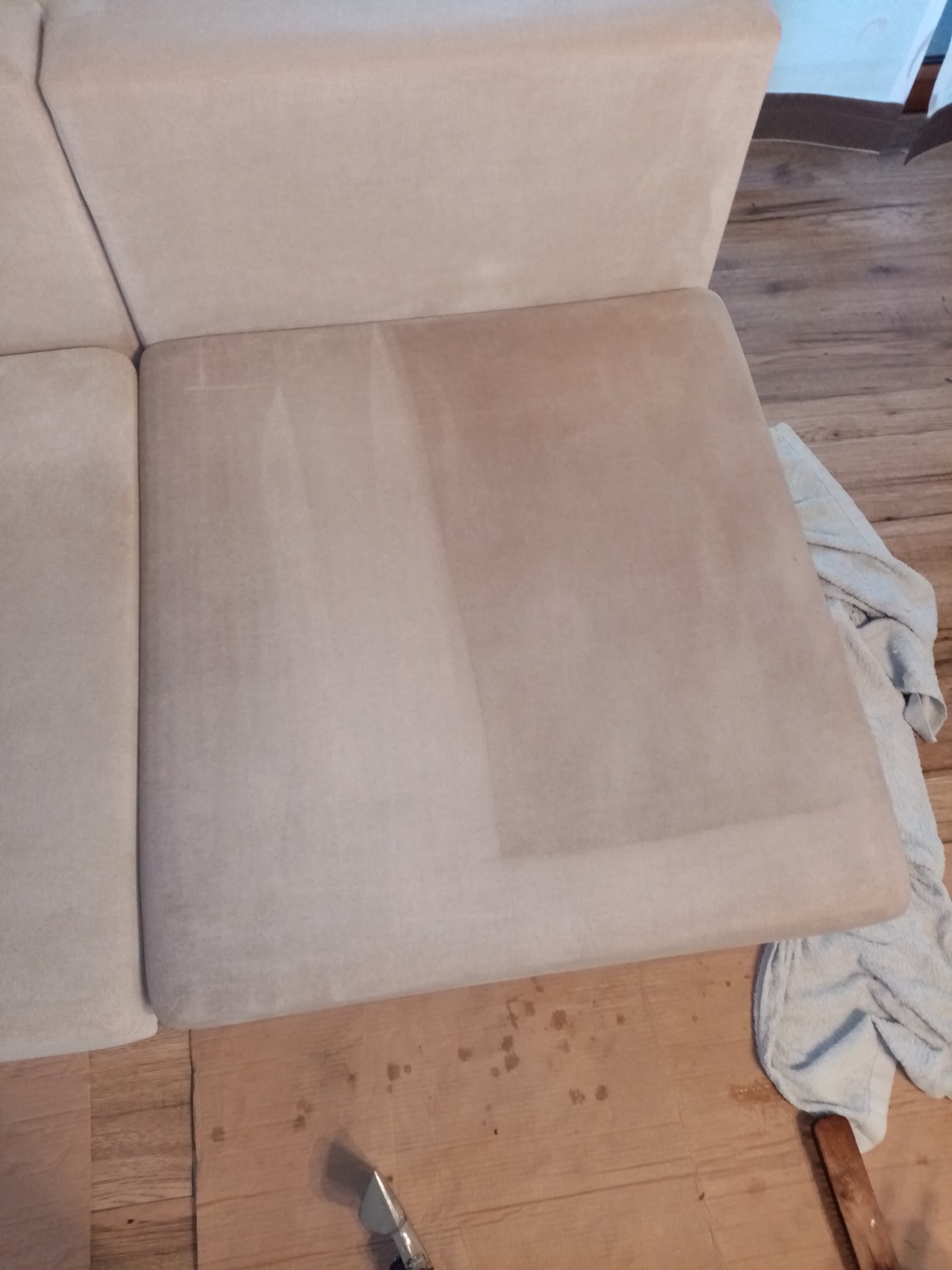 Fragment sofy w trakcie czyszczenia; widoczna wyraźna różnica w kolorze między wyczyszczoną a nieczyszczoną częścią siedziska, obok leży jasny ręcznik i widoczna końcówka odkurzacza piorącego.