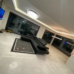 Nowoczesny salon z szarymi płytkami podłogowymi, czarnym narożnikiem i dużymi oknami przesuwnymi, oświetlony listwami LED na suficie i ozdobnym żyrandolem, z parą różowych klapek na dywanie.