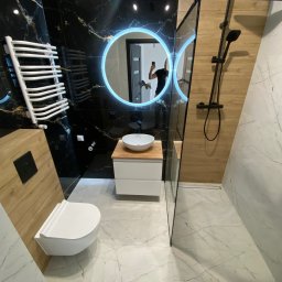 DnL Remonty Budowlano Wykończeniowe - Nowoczesna łazienka z czarnymi płytkami imitującymi marmur ze złotymi żyłami, okrągłym lustrem z podświetleniem LED, białą umywalką nablatową na drewnianym blacie i białą podwieszaną toaletą.