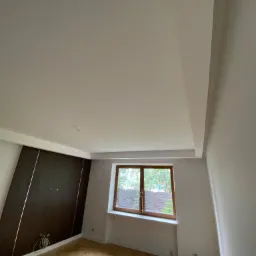 Pokój po remoncie: świeżo pomalowane ściany w odcieniach bieli, drewniane okno z widokiem na zieleń, parkiet w jodełkę i ciemna zabudowa meblowa z metalowymi akcentami.