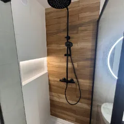 Nowoczesna kabina prysznicowa z czarną armaturą i drewnopodobnymi płytkami, oświetlona wnęka w białej ścianie. Minimalistyczny design.