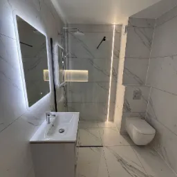 Nowoczesna łazienka z marmurowymi płytkami, podświetlanym lustrem, kabiną prysznicową typu walk-in i podtynkową toaletą. Minimalistyczny design, oświetlenie LED.