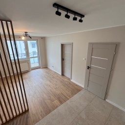 DnL Remonty Budowlano Wykończeniowe - Jasne wnętrze po remoncie: drewnopodobna podłoga, beżowe płytki, dwie szare drzwi z poziomymi wstawkami, drewniana ścianka działowa i oświetlenie sufitowe.