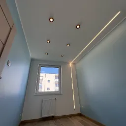 Nowoczesne wnętrze pokoju z błękitnymi ścianami, drewnianą podłogą i oknem. Na suficie zamontowane są punktowe lampy i listwa LED. Widok z dołu, ukazujący detale wykończenia.