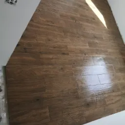 Ułożona podłoga z płytek imitujących drewno w nowym pomieszczeniu z białymi ścianami, widoczna puszka elektryczna bez osprzętu i fragment nierównej podłogi przy ścianie.