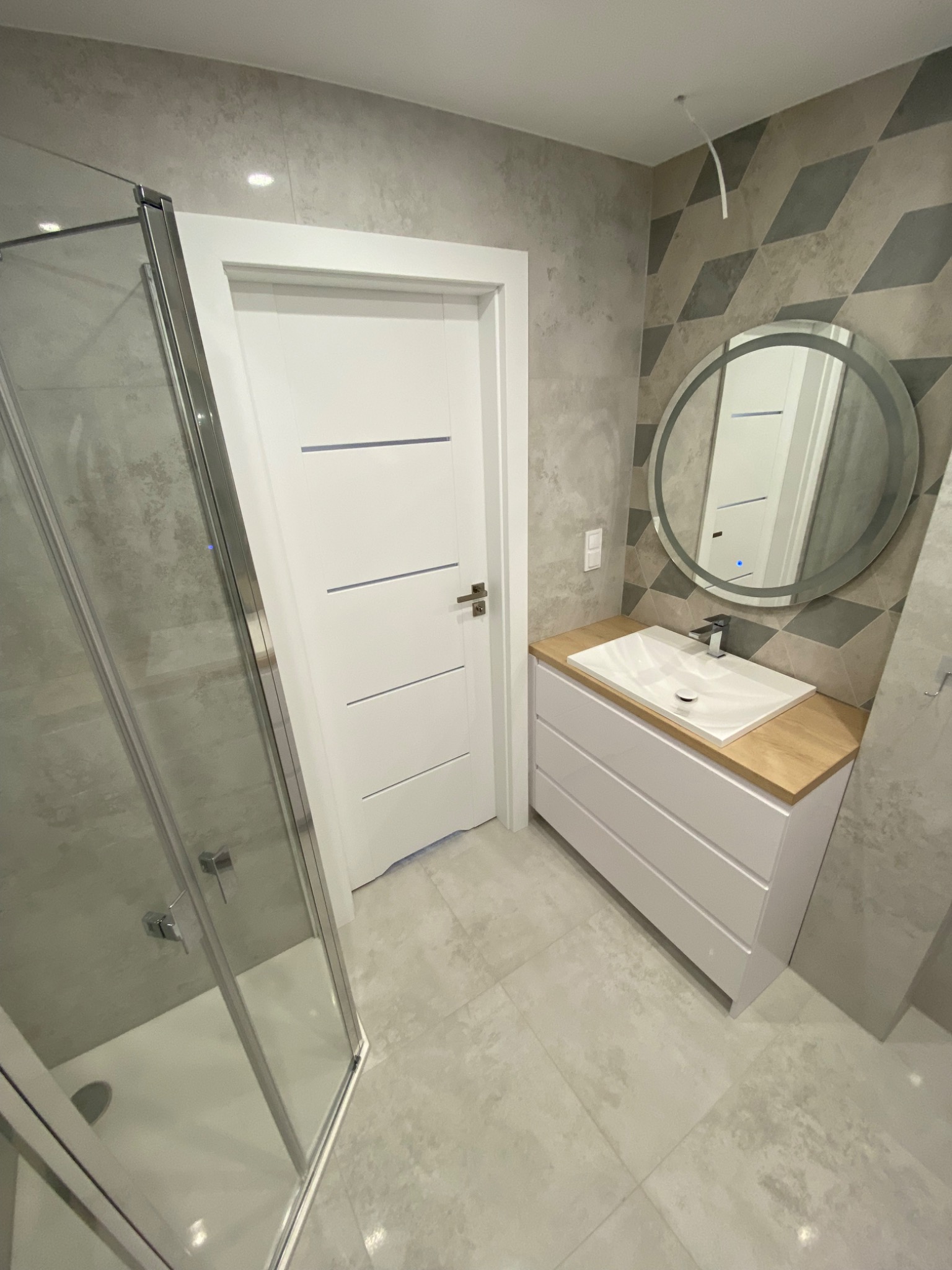 Nowoczesna łazienka z kabiną prysznicową, umywalką nablatową na białej szafce z drewnianym blatem, okrągłym lustrem z podświetleniem LED i geometryczną tapetą na ścianie.