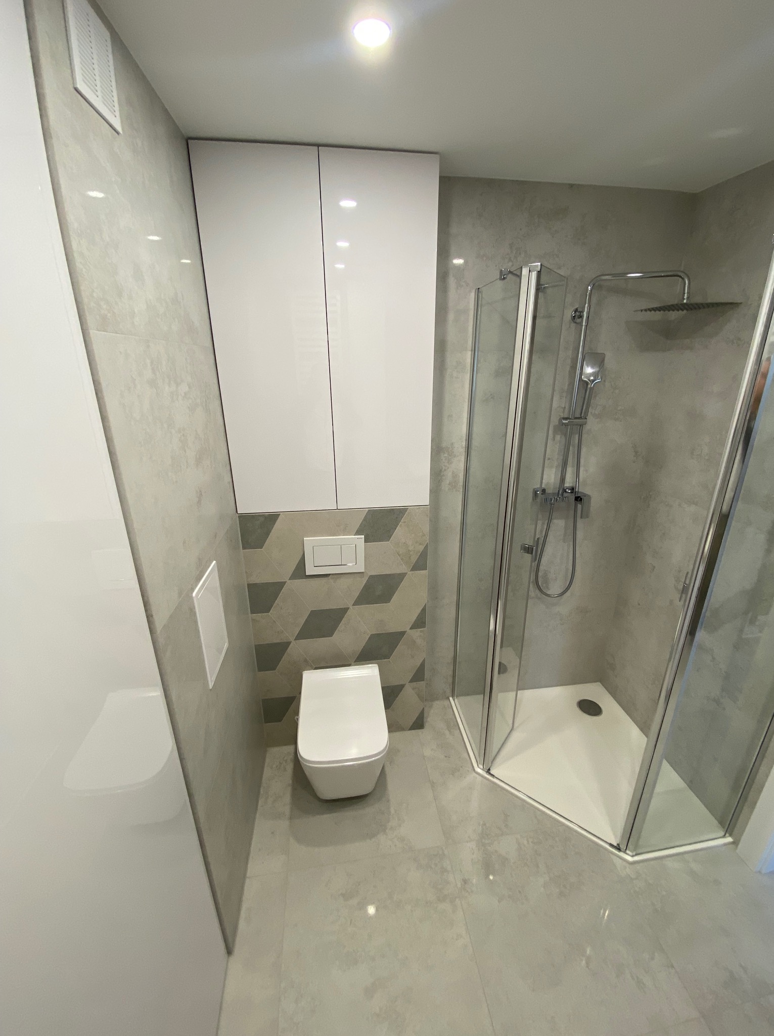 Nowoczesna łazienka po remoncie z kabiną prysznicową narożną, podwieszaną miską WC, szafkami wiszącymi i geometryczną mozaiką nad stelażem.