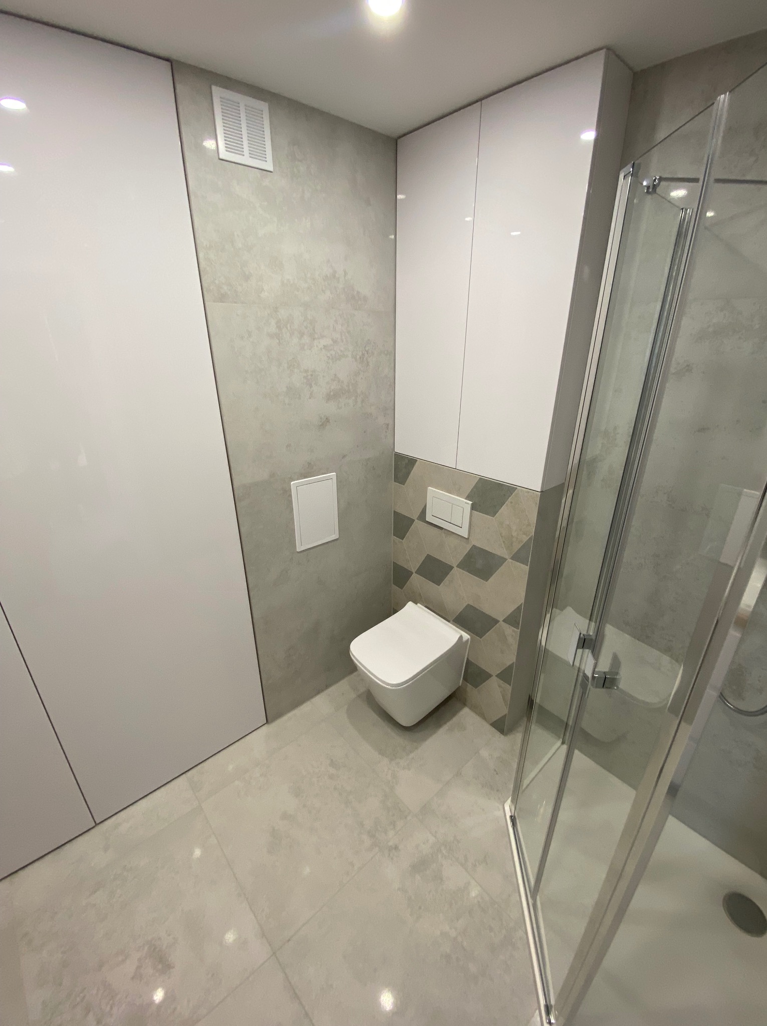 Nowoczesna łazienka po remoncie: szare płytki imitujące beton, biała wisząca toaleta, kabina prysznicowa z transparentnego szkła i białe szafki w zabudowie.