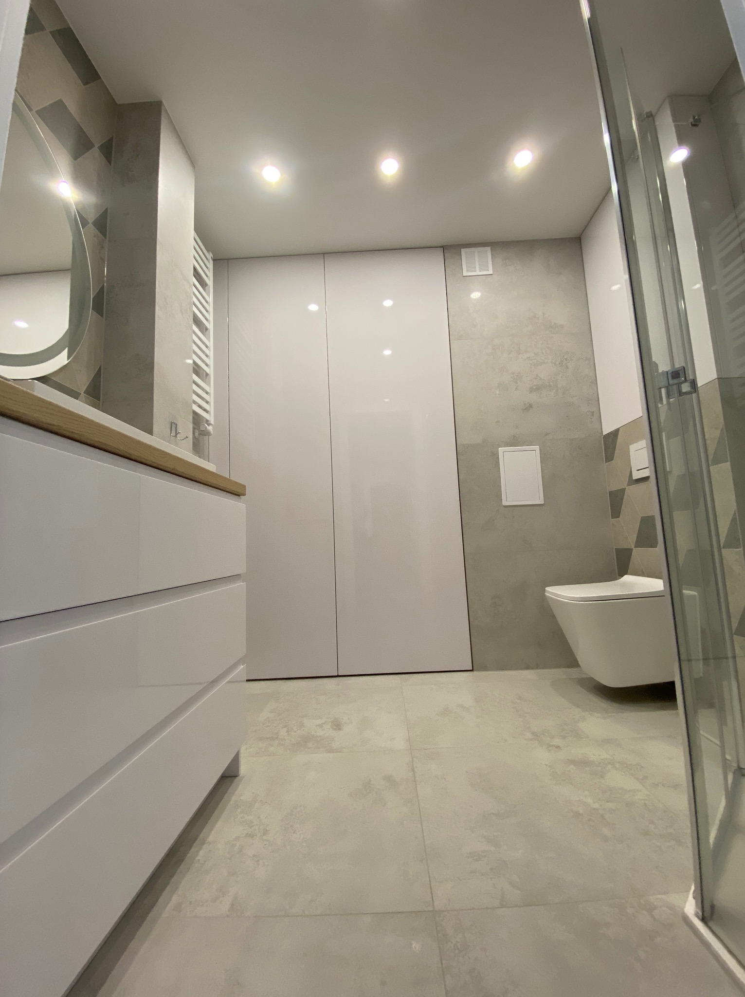 Nowoczesna łazienka z białymi szafkami pod umywalką z drewnianym blatem, podwieszaną toaletą i kabiną prysznicową z widocznymi elementami montażowymi, wykończona płytkami w odcieniach szarości...