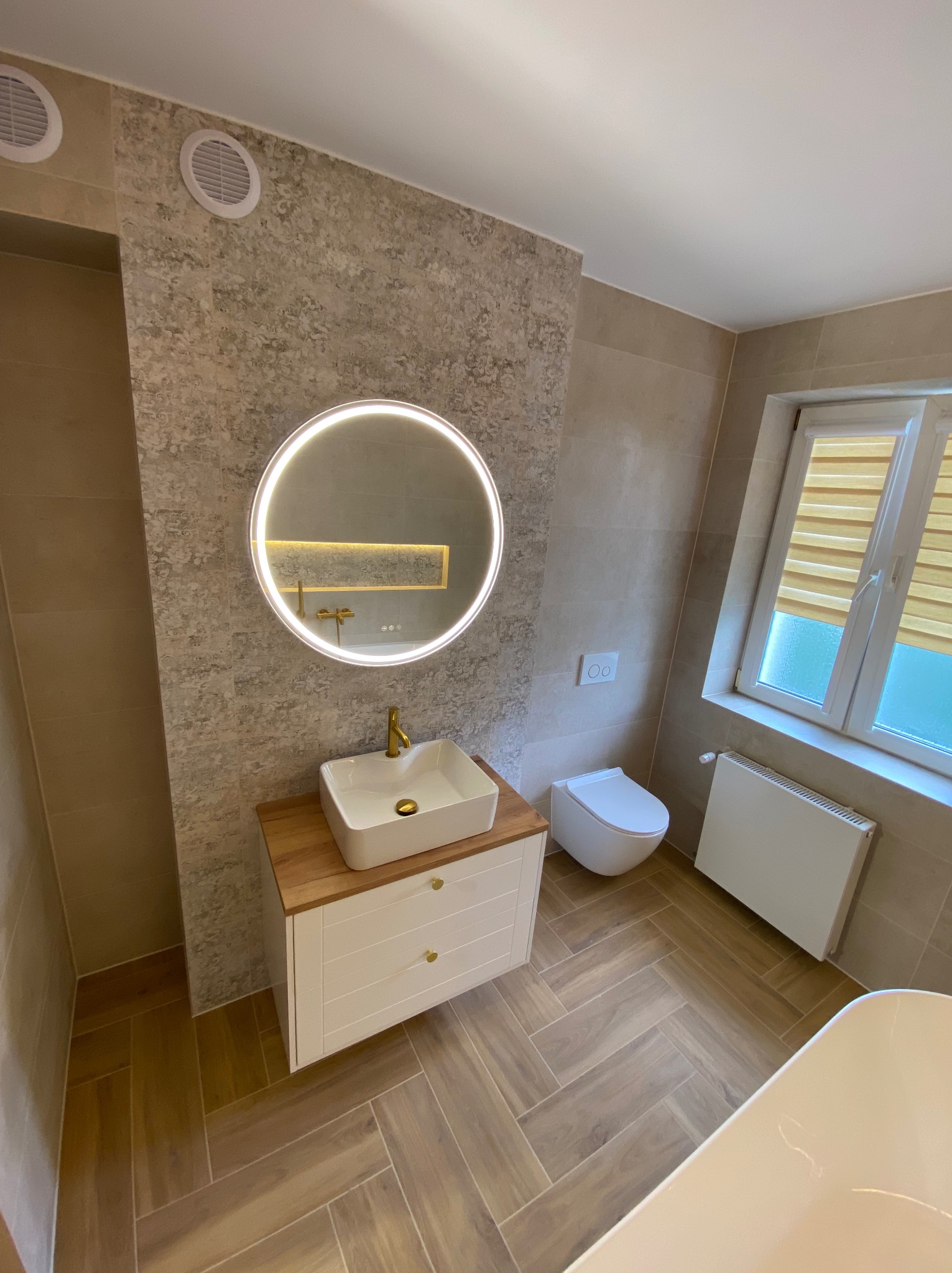 Nowoczesna łazienka z beżowymi płytkami imitującymi kamień, podłoga z paneli ułożonych w jodełkę, okrągłe lustro z podświetleniem LED, biała umywalka nablatowa z złotą baterią i biała szafka...