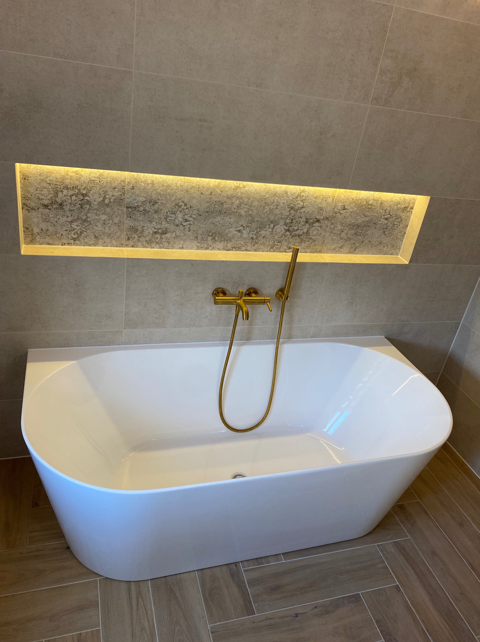 Biała, wolnostojąca wanna z złotą armaturą, umieszczona na podłodze wyłożonej płytkami imitującymi drewno, z podświetlaną wnęką w ścianie wyłożonej szarymi płytkami.
