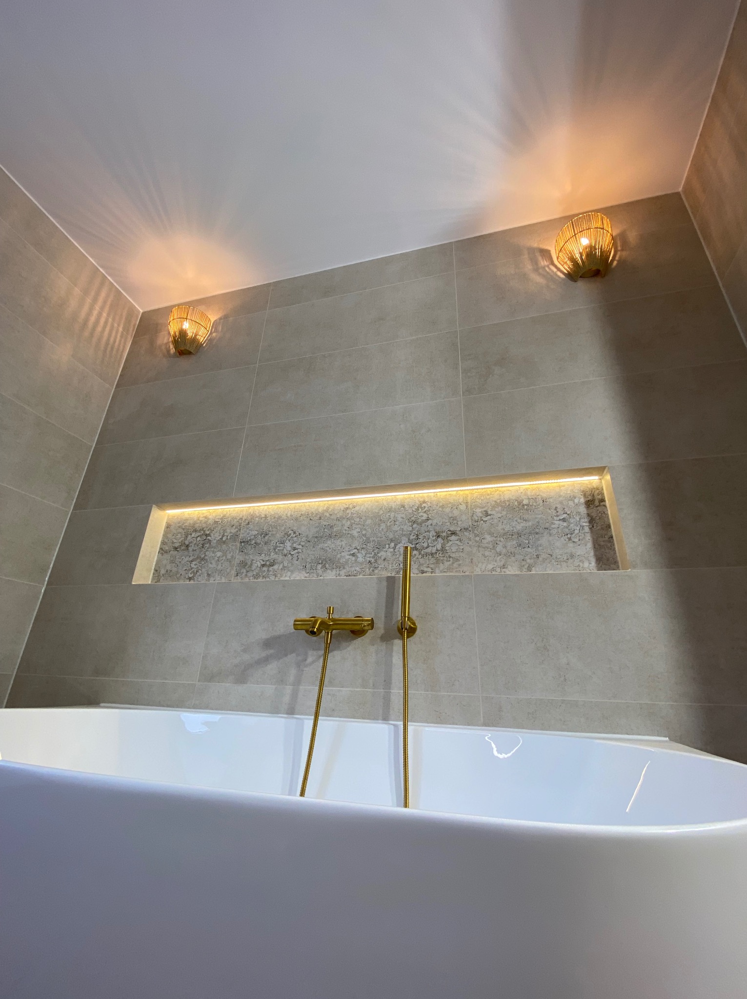 Elegancka łazienka z wanną wolnostojącą, beżowymi płytkami, złotą armaturą i podświetlaną wnęką dekoracyjną w ścianie oraz rattanowymi kinkietami.