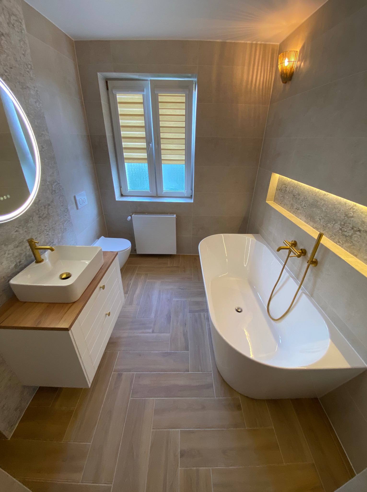 Nowoczesna łazienka z beżowymi płytkami ułożonymi w jodełkę, białą wanną wolnostojącą z baterią w kolorze złotym, umywalką nablatową na szafce z drewnianym blatem i okrągłym lustrem podświetlanym LED.