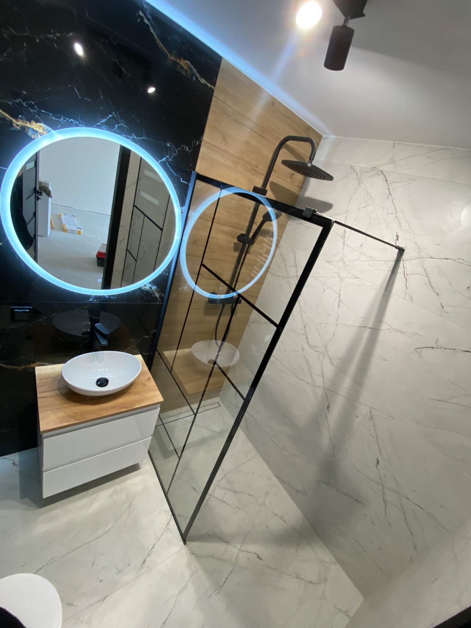 Nowoczesna łazienka z czarnymi marmurowymi płytkami, podświetlanym okrągłym lustrem, drewnianą ścianą i kabiną prysznicową z czarnymi ramami.