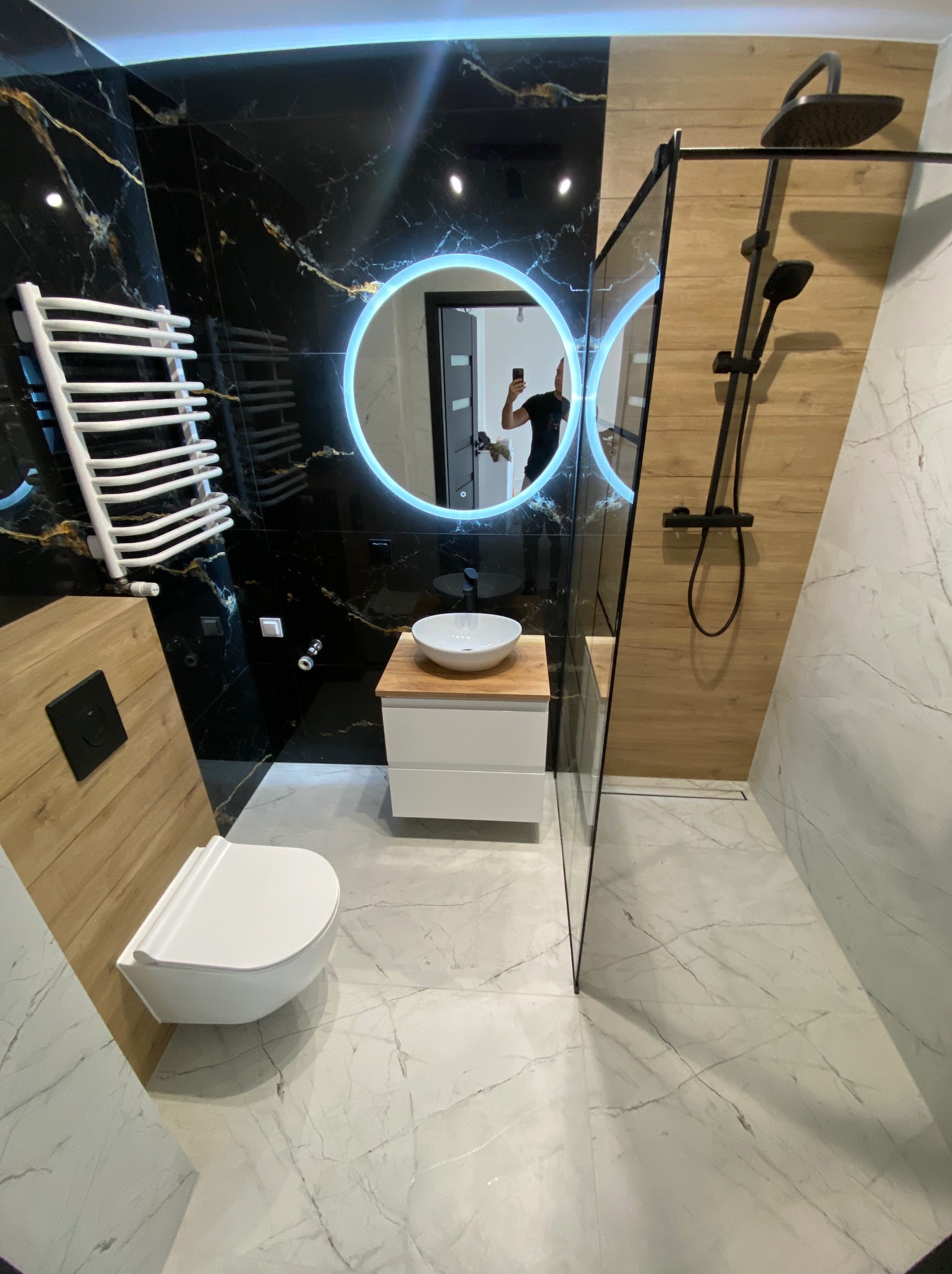 Nowoczesna łazienka z czarnymi płytkami imitującymi marmur ze złotymi żyłami, okrągłym lustrem z podświetleniem LED, białą umywalką nablatową na drewnianym blacie i białą podwieszaną toaletą.