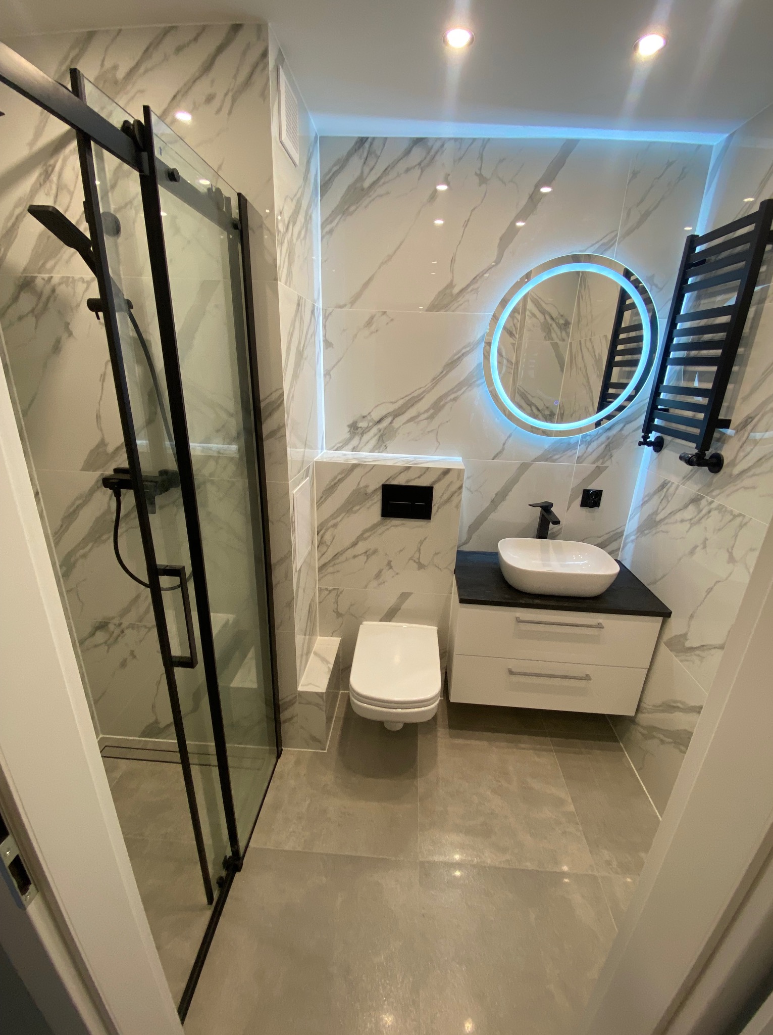 Nowoczesna łazienka z kabiną prysznicową z czarnymi ramami, okrągłym lustrem z podświetleniem LED, umywalką nablatową i grzejnikiem drabinkowym w kolorze czarnym na ścianie wykończonej płytkami...