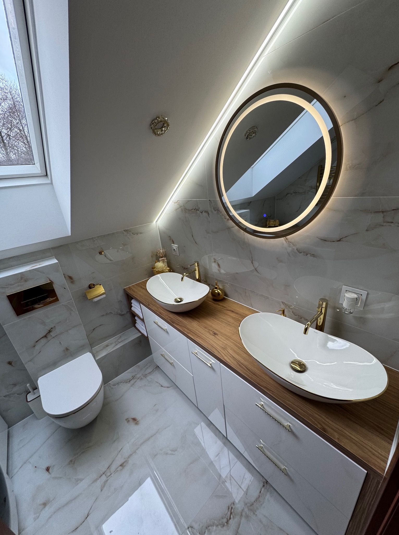 Nowoczesna łazienka na poddaszu z dwoma owalnymi umywalkami, okrągłym lustrem z podświetleniem LED, białą szafką z drewnianym blatem i marmurowymi płytkami na ścianach oraz podłodze.
