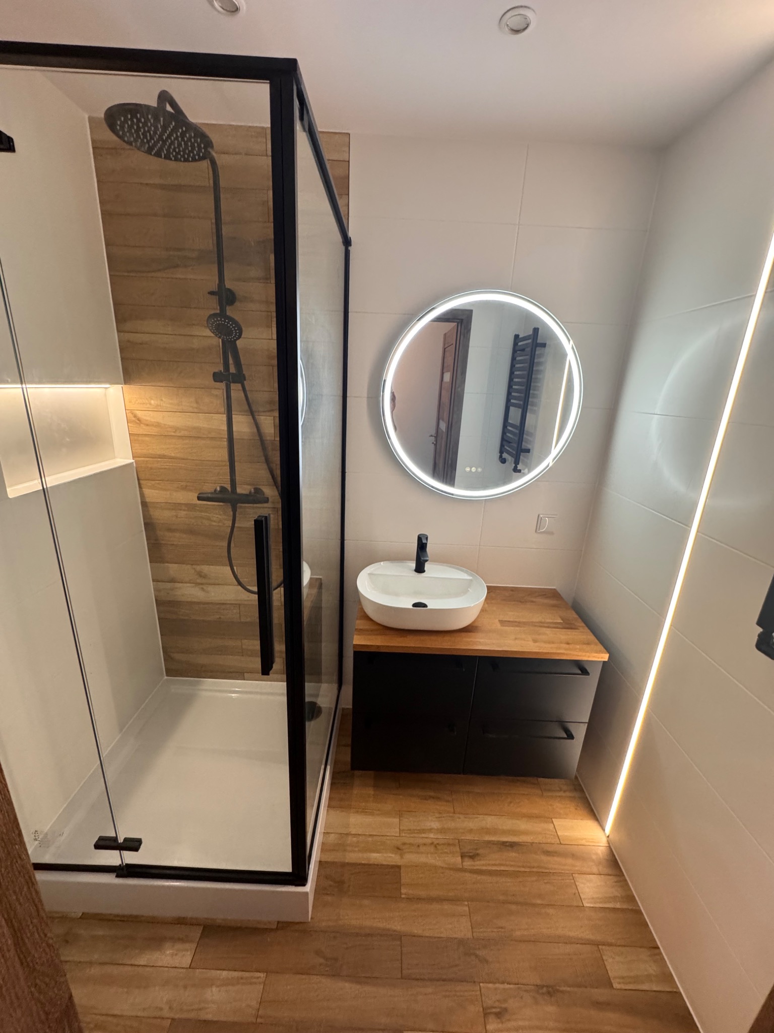 Nowoczesna łazienka z kabiną prysznicową typu walk-in, okrągłym lustrem z podświetleniem LED i drewnopodobną podłogą. Minimalistyczny design z czarnymi akcentami.