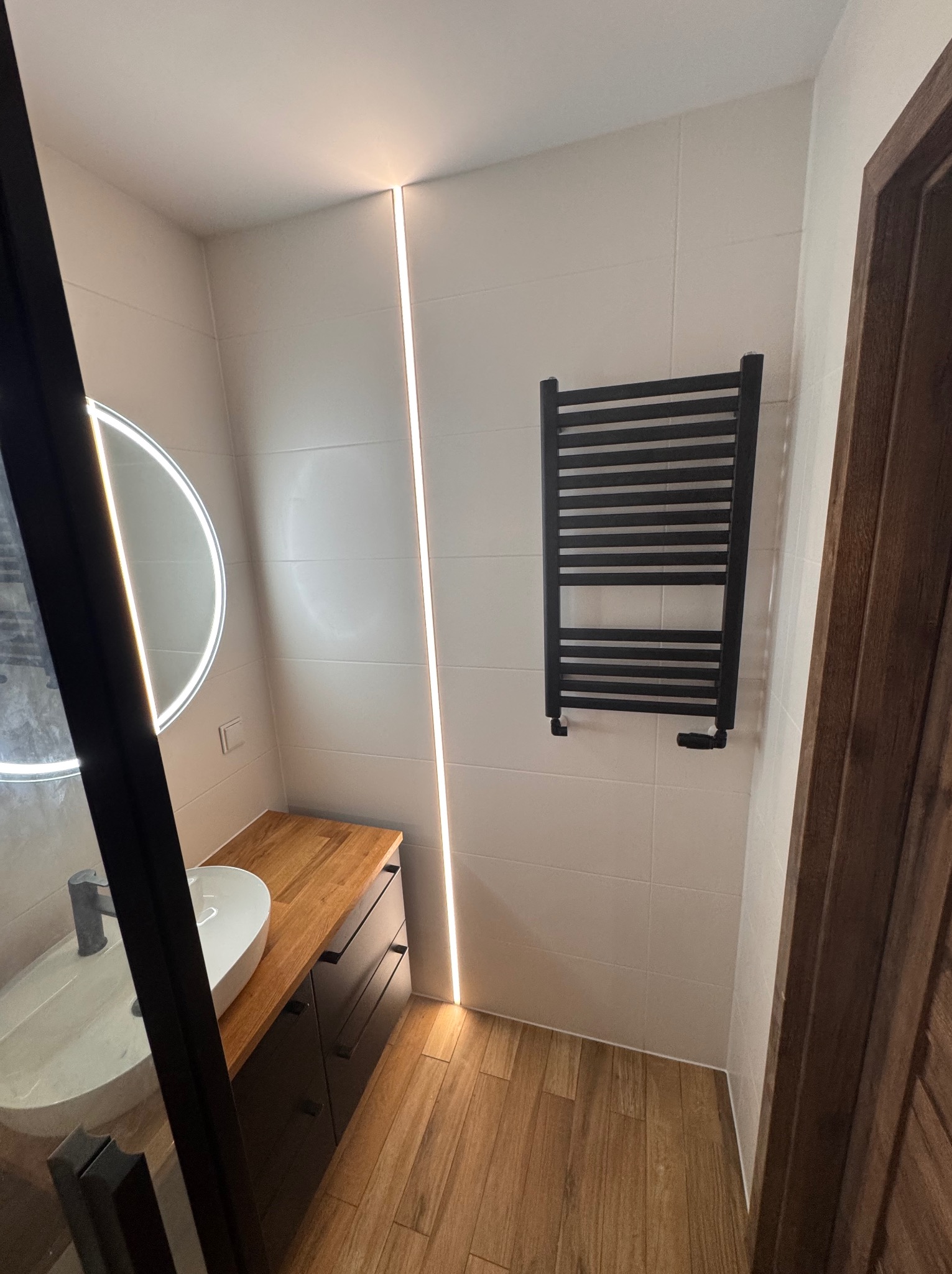 Nowoczesna łazienka z drewnianą podłogą i blatem, białymi płytkami, podświetlanym lustrem i grzejnikiem drabinkowym. Minimalistyczny design z akcentami ciepłego drewna.