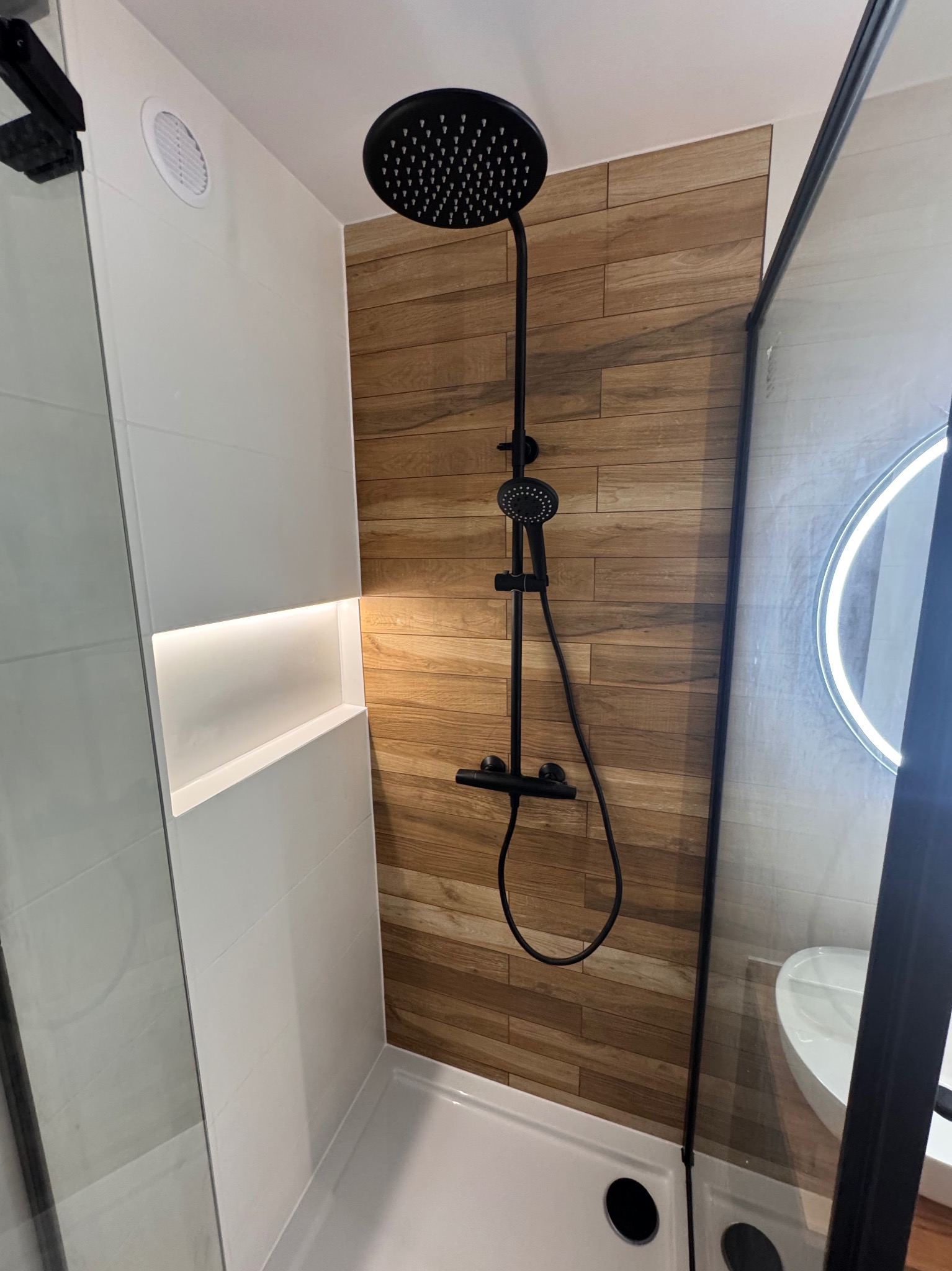 Nowoczesna kabina prysznicowa z czarną armaturą i drewnopodobnymi płytkami, oświetlona wnęka w białej ścianie. Minimalistyczny design.